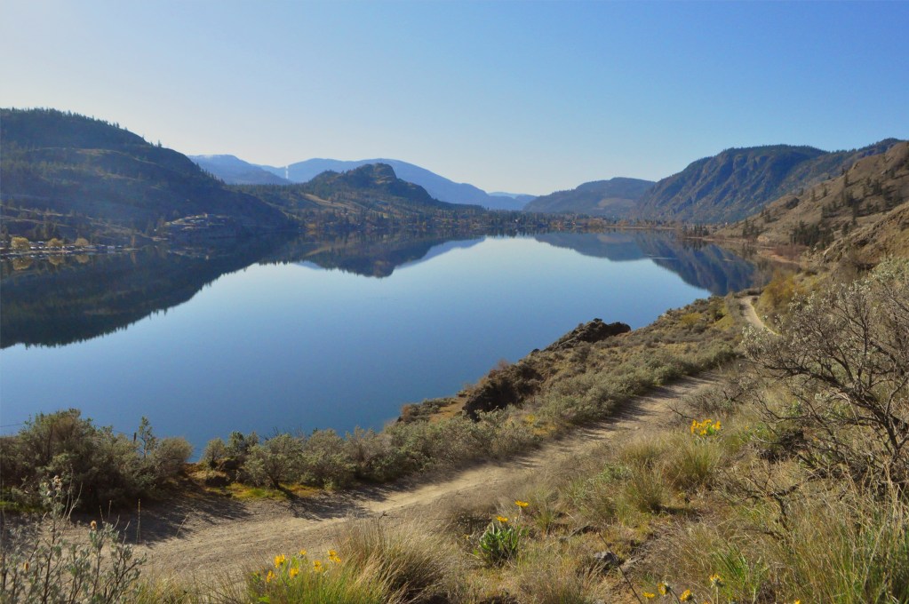 a still Skaha Lake on a clear sunny day with mountains in the foreground and background,