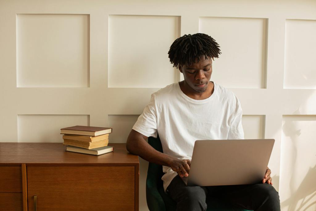 a young man with dreads wearing a white t-shiirt sitting next to a desk with a laptop on his lap
